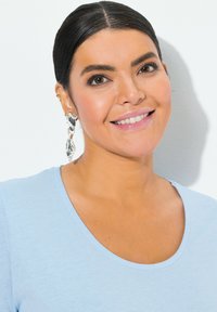 Stříbrné točené náušnice, světle modré tričko s výstřihem do lodičky, hladký materiál, měkká tkanina, přirozený make-up a tmavé vlasy stažené dozadu.
