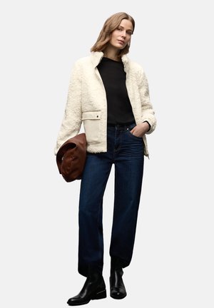 Kremowa kurtka z sztucznego baranka z suwakiem, czarny top, ciemnoniebieskie proste jeansy i czarne botki, trzymająca brązową torbę.