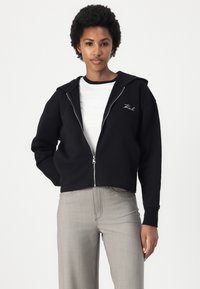 Schwarze, verkürzte Zip-Hoodie mit Kapuze und gesticktem Logo. Kombiniert mit grauen, geraden Hosen und einem weißen T-Shirt darunter.