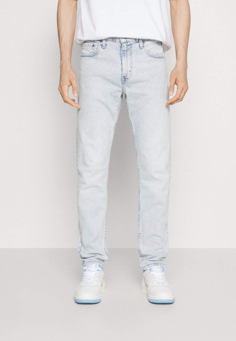 Homme portant un jean bleu clair, des baskets blanches et un T-shirt blanc rentré dans le pantalon, debout devant un fond clair uni.