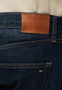 Σκούρο μπλε τζιν με πορτοκαλί ραφές, καφέ δερμάτινο ταμπελάκι με την επιγραφή «Tommy Jeans» και ένα μικρό λογότυπο σε κόκκινο, άσπρο και μπλε στην πίσω τσέπη.