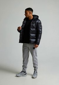 Melna pūkaina jaka ar spīdīgu tekstūru un logotipa detaļu, kombinēta ar pelēkām sporta biksēm un pelēkām sporta kurpēm, noturēta ar mikrofonu.