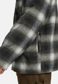 Close-up van de arm van een persoon die een geruite flanellen shirt draagt in blauwe, witte en bruine tinten, met een lichte focus op de textuur en het patroon van de mouw.