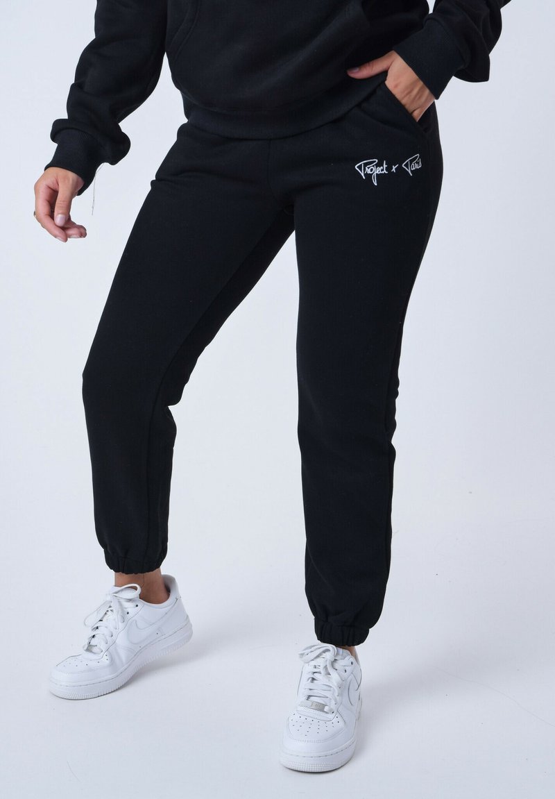Zwarte sportbroeken met een elastische tailleband en boorden, met een wit logo op de bovenbeen. Draagt witte sportschoenen.