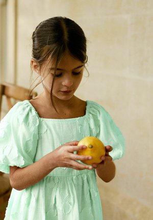 Dijete drži žuti citrus, obučeno u haljinu mint zelene boje s teksturom, s napuhanim rukavima i kvadratnim izrezom. Pozadina je neutralna.
