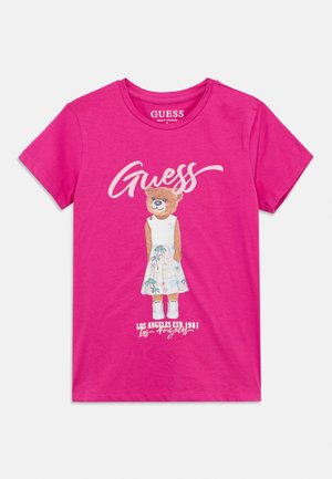 Tricou roz aprins cu un urs purtând un top alb, fustă cu model de palmier, adidași și textul „Guess” și „Los Angeles Est. 1981”.