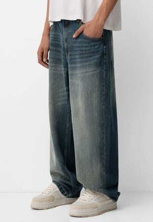 Personne portant un jean bleu délavé à jambes larges avec la main dans la poche et des baskets blanches, debout devant un fond blanc uni.