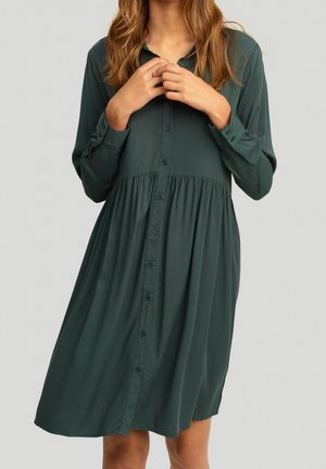 Langärmliges, knöpfbares Kleid in Dunkelgrün. Hergestellt aus weichem Stoff mit einer gerafften Taille und einer lockeren Passform, ausgestattet mit einem klassischen Kragen.