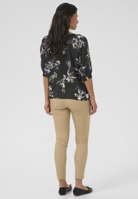 Blusa nera a fiori con maniche a tre quarti, caratterizzata da un vivace motivo floreale in viola e giallo, abbinata a pantaloni beige aderenti.