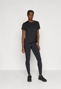 Woman standing against wall wearing black ruched side t-shirt, black leggings, and black athletic shoes.