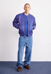 Chaqueta bomber morada con cremallera, camiseta azul debajo, jeans de mezclilla azul holgados y zapatos marrones con acentos de mocasín sobre un fondo liso.