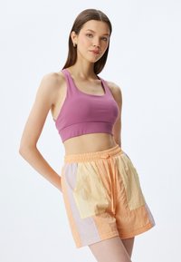 Sujeción deportiva de color lila claro combinada con pantalones cortos de parcheo en tonos durazno y pastel, con cintura elástica y bolsillos laterales, de textura suave.