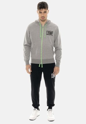 Giovane uomo che indossa felpa con cappuccio grigia con cerniera e cordini verdi e pantaloni della tuta neri con scarpe da ginnastica bianche, in piedi di fronte.