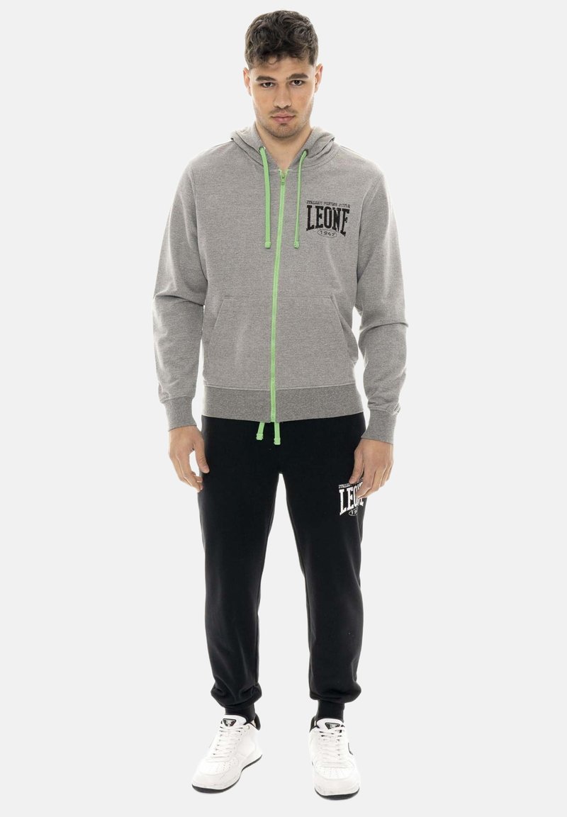 Jeune homme portant un sweat à capuche zippé gris avec des cordons verts, un pantalon de jogging noir et des baskets blanches, debout face à nous.