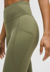 Hochtaillierte grüne Sportleggings aus glattem Material, mit geschwungenen Nähten, Seitentaschen und kleinen reflektierenden Akzenten.