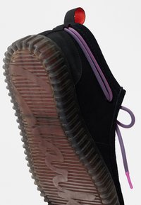 Μαύρο αθλητικό παπούτσι με διάφανη ραβδωτή σόλα, ροζ και μωβ κορδόνια, και κόκκινη λαβή σε λευκό φόντο.