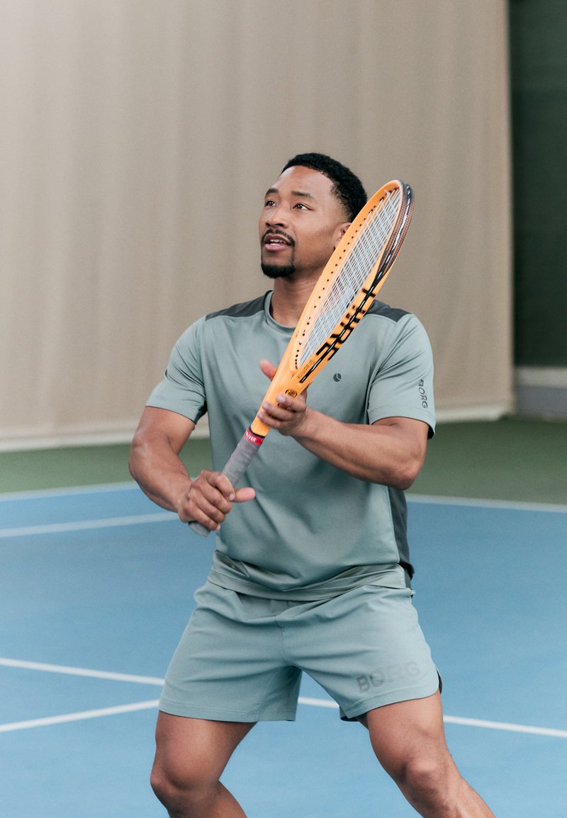 Atleta com um fato desportivo verde claro, segurando uma raquete de ténis laranja, prepara-se para acertar na bola num court azul com um fundo bege.