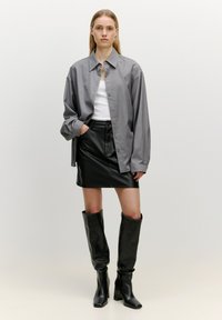 Graues, übergroßes Hemd mit Knöpfen, weißes Tanktop, schwarze Leder-Minirock und hohe schwarze chunky Stiefel, stehend vor einem neutralen Hintergrund.