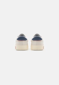 Zapatillas blancas con parte superior de cuero suave, un tirador del talón de color azul oscuro y suela de goma color crema. Diseño minimalista con acentos en contraste.