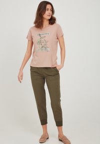 Camiseta rosa claro con un gráfico de planta y texto, combinada con pantalones verde oliva ajustados que presentan puños elásticos y bolsillos frontales.