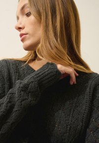 Tmavě šedý pletený svetr s texturovaným vzorem, který má žebrované manžety a výstřih do V. Měkká, silná tkanina je viditelná zblízka.