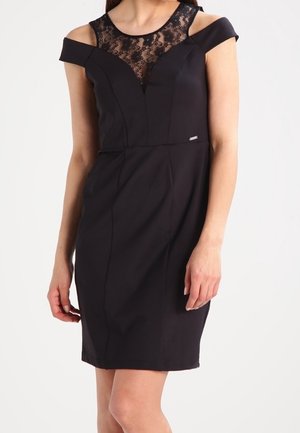 Frau trägt ein schwarzes knielanges Kleid mit schulterfreien Trägern und einem Spitzenbesatz am Ausschnitt, steht vor einem schlichten Hintergrund.