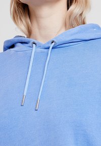 Sudadera con capucha azul claro con cordones de puntas metálicas, tela texturizada y un ajuste relajado con capucha. Sin patrones ni detalles adicionales.