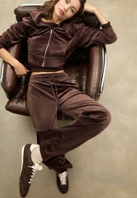Brązowy welurowy krótki hoodie z przednią kieszenią, w zestawie z dopasowanymi dresami. Model nosi białe i brązowe sneakersy z kremowymi skarpetami.