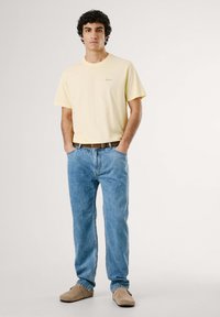 Camiseta de algodón amarilla claro con mangas cortas, jeans de mezclilla azul y zapatos tipo slip-on beige. Diseño sencillo con un corte relajado y sin patrones prominentes.