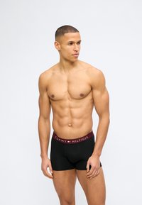 Γυμνασμένος άνδρας χωρίς πουκάμισο που φοράει μαύρα μποξεράκια Tommy Hilfiger με μπορντό ελαστική μέση, στέκεται μπροστά σε απλό λευκό φόντο.