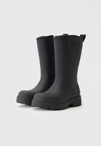 Bottes en caoutchouc noires avec une semelle plate, un bout rond et un talon légèrement surélevé. Présente un tirant à l'arrière et une texture matte lisse.