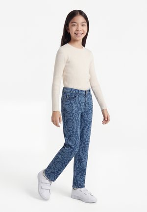 Nasmejana deklica stoji, oblečena v kremasto rebrast dolg rokav, modre kavbojke z vzorcem in bele superge z ježki.