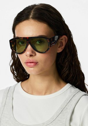Jeune femme aux cheveux ondulés et foncés portant de grandes lunettes de soleil écaille avec des verres jaunes et un haut blanc à plusieurs couches.