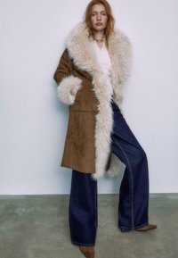 Brun ruskind frakke med en stor, cremefarvet kunstpelskrave og -manchetter, parret med mørke, vide jeans og brune ankelstøvler på en grå gulvbaggrund.