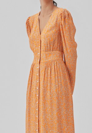 Femme portant une robe chemise orange à manches longues avec des motifs floraux blancs et une taille cintrée, sur un fond uni.