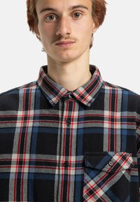 Koszula w kratę z guzikami, w kolorze czarnym, niebieskim, czerwonym i białym. Miękka tkanina, z kieszonką na piersi i zaokrąglonym kołnierzykiem.