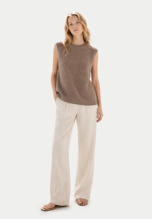 Femme portant un haut en tricot marron sans manches, un pantalon beige ample et des sandales, debout avec une main dans la poche devant un fond uni.