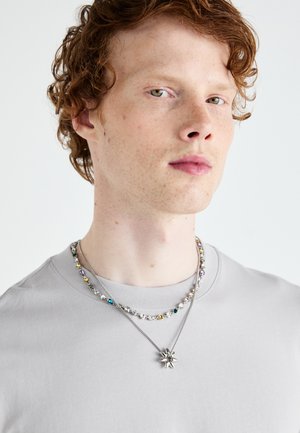 Sidabrinės ir spalvotos karoliukų kolje, sluoksniuojamos ant paprasto pilko marškinėlių. Kolje turi žvaigždės formos pakabuką ir įvairių tekstūrų karoliukus.