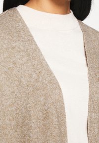 Béžový pletený cardigan s otevřeným předním dílem, měkká textura a bílé vnitřní svršek s žebrováním. Materiál se zdá být teplý a pohodlný, vhodný pro vrstvení.