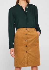Tummanvihreä pitkähihaine paita, jossa on kaulus ja kaksi nappia. Vaaleanruskea corduroy-hame, jossa on etunappikiinnitys ja taskut, ja joka on varustettu ribboituneella pinnalla.