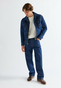 Τζιν μπουφάν και παντελόνι σε σκούρο μπλε με πορτοκαλί ραφές. Το πουκάμισο είναι ανοιχτό μπεζ. Καφέ παπούτσια με υφή στη σόλα. Άνετη εφαρμογή.