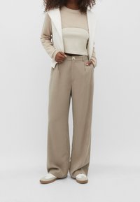 Beżowy krótki sweter noszony pod białą bezrękawnik, w zestawie z szerokimi beżowymi spodniami i białymi sneakersami z brązowymi podeszwami.