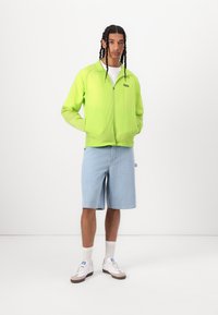 Neon green zip-up jacket with two front pockets, paired with light blue textured shorts and white sneakers. Plain white T-shirt underneath.