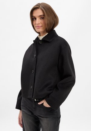 Schwarze, kurze Jacke aus strukturiertem Stoff mit kragenförmigem Ausschnitt, großen Knöpfen und weiten Ärmeln. Kombiniert mit dunklen Jeans.