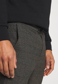 Pantalones de cuadros grises confeccionados en una tela texturada, con sutiles líneas naranjas, pliegues y cintura con cordón. Top negro de manga larga visible.