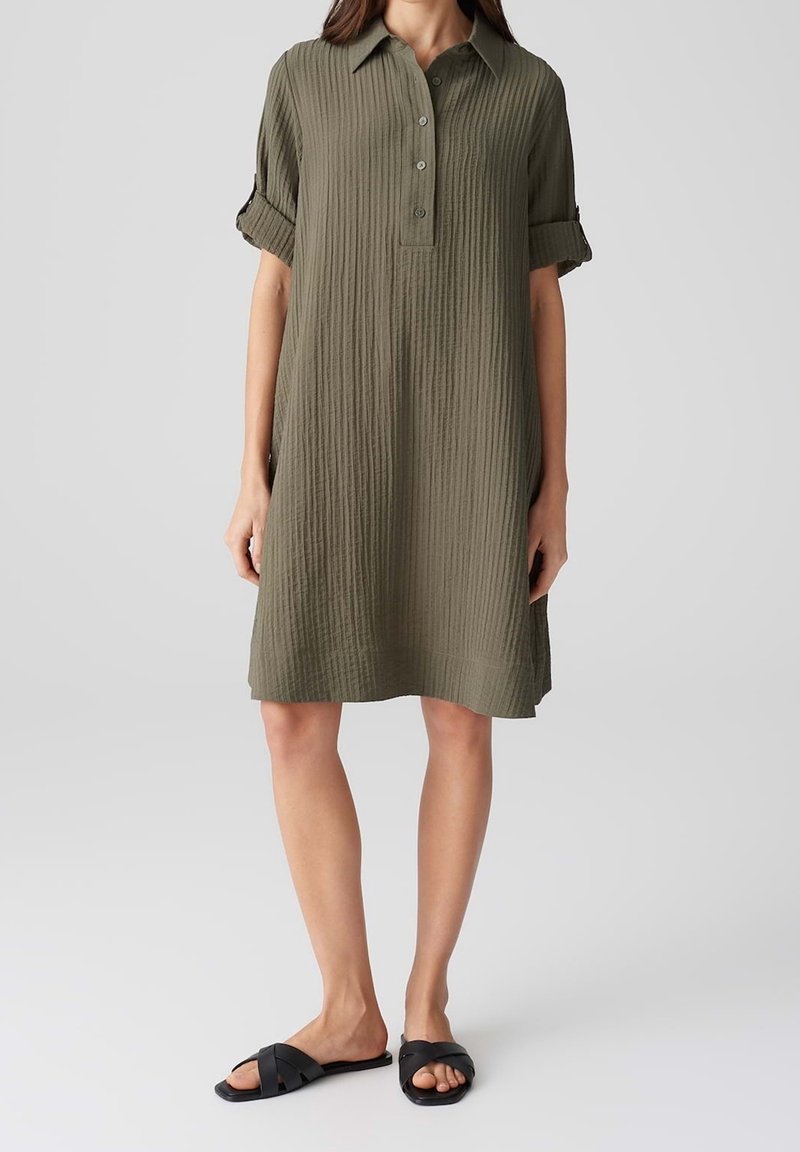 Femme portant une robe chemise texturée longueur genoux de couleur vert olive, manches retroussées, et des sandales noires à enfiler sur un fond gris clair.