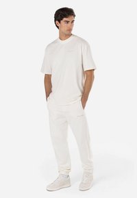 Camiseta de algodón color crema y joggers a juego con puños acanalados, que presentan un logo sutil en el lado izquierdo del pecho y en el muslo. Las zapatillas completan el look.