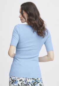 Vrouw met golvend bruin haar, gekleed in een lichtblauw, aansluitend ribbelshirt en bloemenpatroon broek, met haar rug naar de camera toe, op een witte achtergrond.