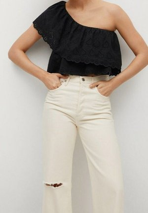 Mujer con top corto negro de un solo hombro con volantes y vaqueros de talle alto color marfil roto con un desgarro sobre la rodilla, manos en los bolsillos.