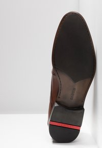 Brun lædersko med spids tå, slankt design og en kontrastfyldt mørk gummisål. Har rød accent på hælen og præget branding.